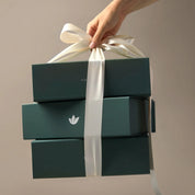 Three stacked dark teal gift boxes tied with a wide white ribbon, held by a person's hand against a plain warm beige background.