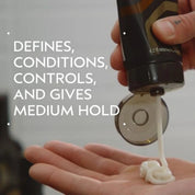 A hand squeezes a dark brown hair product tube to dispense white cream into another hand, with large white text on the left reading 'DEFINES, CONDITIONS, CONTROLS, AND GIVES MEDIUM HOLD'.