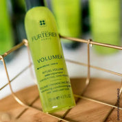 A bottle of Rene Furterer Paris Volumea Volumizing Conditioning Spray, in a translucent yellow-green color, resting on a gold wire tray over a wooden surface with a blurred green background.
