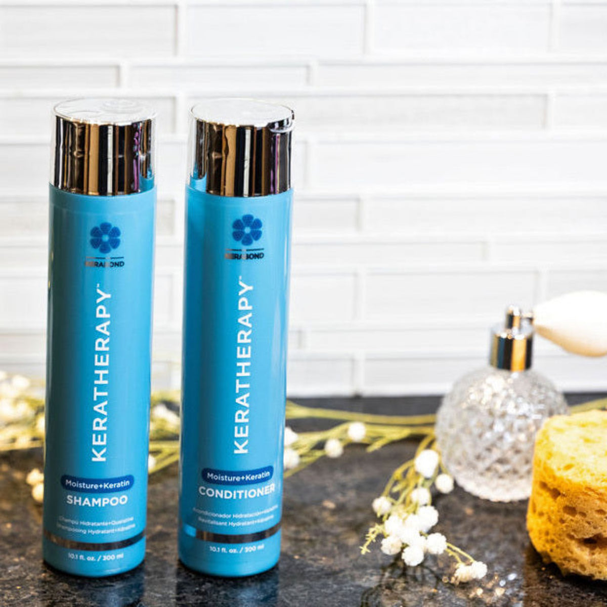 Two blue Keratherapy shampoo and conditioner bottles with shiny silver caps, displayed on a dark countertop with decorative flowers, a sponge, and a glass bottle against a white subway tile wall.