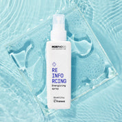 A white bottle of Morphosis Reinfoicing Energizing spray stands upright on a clear square glass tile over a light blue water-textured background.