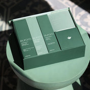 A boxed skincare set from eurora, featuring three tall rectangular boxes and one square box in matching sage green packaging, displayed on a round pedestal with dramatic shadow patterns.