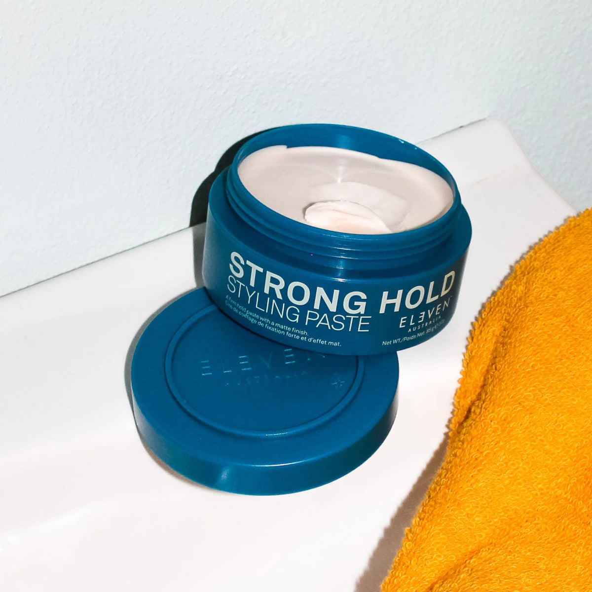 Blue jar of 'Strong Hold Styling Paste' with a yellow towel on a white surface