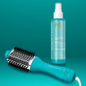 Moroccanoil Frizz Shield Spray bottle and a turquoise round brush styling tool on a teal gradient background.