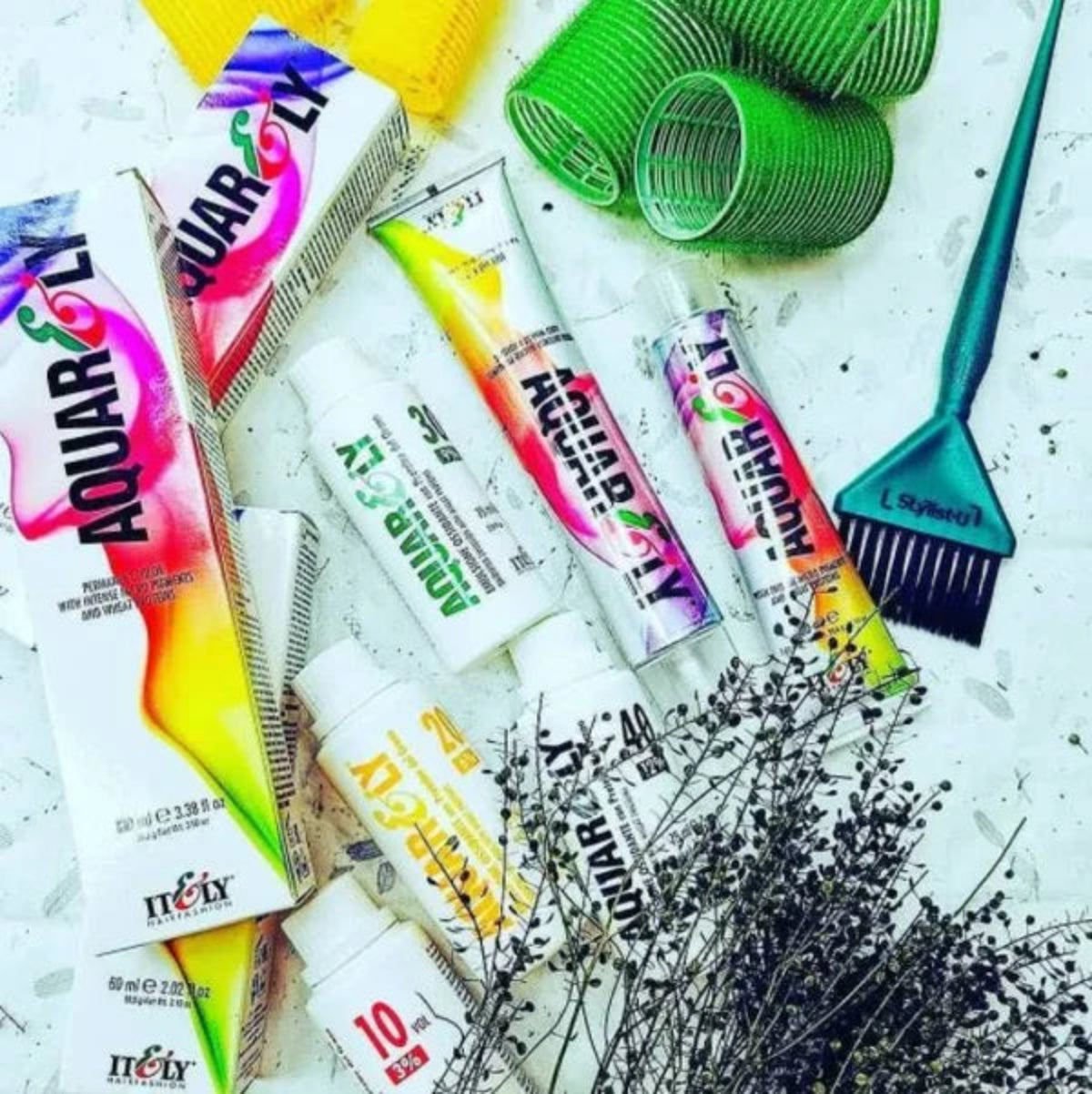 A styled flat-lay of hair dye products from the brand NULY AQUA RLY, including several white squeeze tubes with colorful labels, matching product boxes, a teal hair applicator brush, green plastic curlers, and dried lavender sprigs on a white textured background.